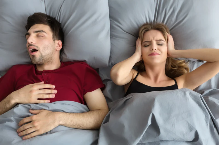 Man snoring in bed while the woman beside him covers her ears