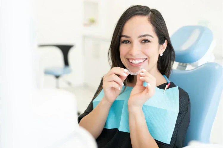 A woman at a dentist's office, holding an aligner and smiling.